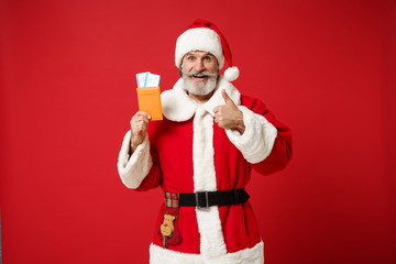 Fototapeta na wymiar Elderly gray-haired mustache bearded Santa man in Christmas hat isolated on red background. New Year 2020 celebration holiday concept. Mock up copy space. Holding passport ticket showing thumb up.