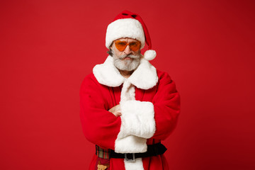 Serious Elderly gray-haired mustache bearded Santa man in Christmas hat, sunglasses posing isolated on red background. Happy New Year 2020 celebration concept. Mock up copy space. Hold hands crossed.