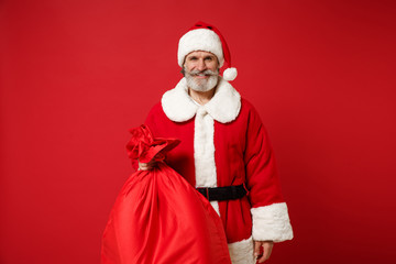Fototapeta na wymiar Smiling elderly gray-haired mustache bearded Santa man in Christmas hat posing isolated on red background. Happy New Year 2020 celebration holiday concept. Mock up copy space. Hold bag with presents.