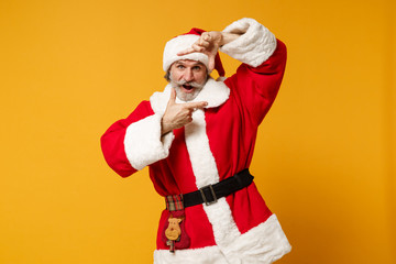 Fototapeta na wymiar Cheerful elderly gray-haired mustache bearded Santa man in Christmas hat posing isolated on yellow background. New Year 2020 celebration concept. Mock up copy space. Making hands photo frame gesture.