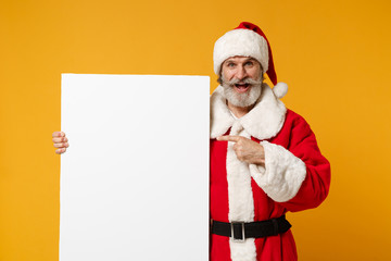 Elderly gray-haired mustache bearded Santa man in Christmas hat posing isolated on yellow background. New Year 2020 celebration concept. Mock up copy space. Pointing finger on white empty billboard.