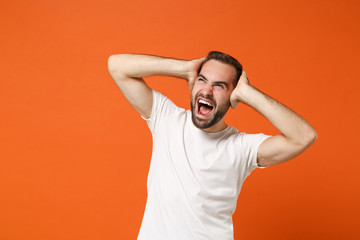 Fototapeta na wymiar Frustrated young man in casual white t-shirt posing isolated on orange wall background, studio portrait. People lifestyle concept. Mock up copy space. Screaming, looking up, putting hands on head.