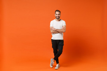 Obraz na płótnie Canvas Smiling attractive young man in casual white t-shirt posing isolated on orange wall background studio portrait. People sincere emotions lifestyle concept. Mock up copy space. Holding hands crossed.