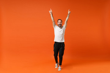 Funny young man in casual white t-shirt posing isolated on orange background, studio portrait. People sincere emotions lifestyle concept. Mock up copy space. Rising hands, pointing index fingers up.