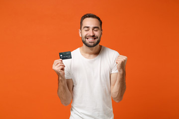 Happy young man in casual white t-shirt posing isolated on orange wall background. People lifestyle concept. Mock up copy space. Holding credit bank card, doing winner gesture, keeping eyes closed.