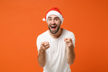 Fototapeta premium Excited young Santa man in white t-shirt, Christmas hat posing isolated on orange background. Happy New Year 2020 celebration holiday concept. Mock up copy space. Pointing index fingers on camera.