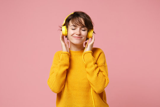 Relaxed Young Brunette Woman Girl In Yellow Sweater Posing Isolated On Pastel Pink Background In Studio. People Lifestyle Concept. Mock Up Copy Space. Listen Music With Headphones Keeping Eyes Closed.