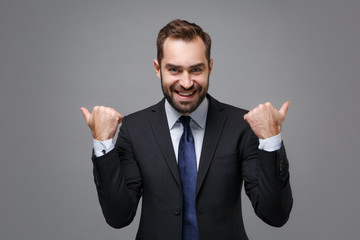 Cheerful young bearded business man in classic black suit shirt tie posing isolated on grey background in studio. Achievement career wealth business concept. Mock up copy space. Pointing thumbs aside.