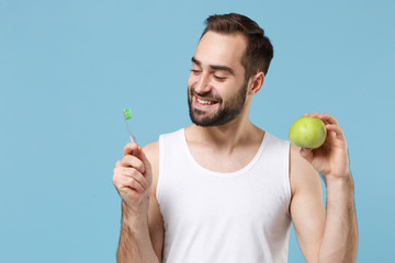 Bearded young man 20s years old in white shirt hold brush and green apple isolated on blue pastel wall background studio portrait. Skin care healthcare cosmetic procedures concept Mock up copy space.