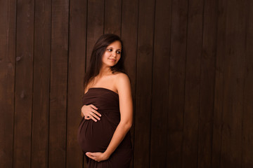 Fototapeta premium Pregnant woman in brown dress holds hands on belly on a dark brown background. Pregnancy, maternity, expectation concept. Beautiful tender mood photo of pregnancy