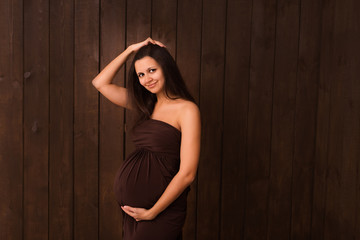 Fototapeta premium Pregnant woman in brown dress holds hands on belly on a dark brown background. Pregnancy, maternity, expectation concept. Beautiful tender mood photo of pregnancy