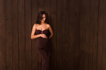 Naklejka premium Pregnant woman in brown dress holds hands on belly on a dark brown background. Pregnancy, maternity, expectation concept. Beautiful tender mood photo of pregnancy