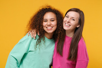 Fototapeta premium Two smiling women friends european and african american girls in pink green clothes posing isolated on yellow orange background studio portrait. People lifestyle concept. Mock up copy space. Hugging.