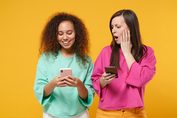 Two shocked women friends european african american girls in pink green clothes posing isolated on yellow background. People lifestyle concept. Mock up copy space. Using mobile phone, typing message.