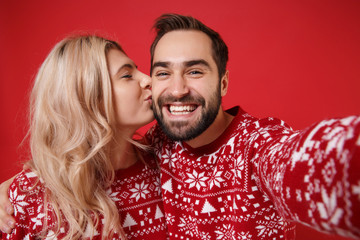 Close up of merry young couple guy girl in Christmas sweaters posing isolated on red background. Happy New Year 2020 celebration party concept. Mock up copy space. Doing selfie shot on mobile phone.