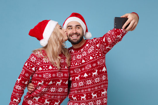 Pretty young couple guy girl in Christmas sweater Santa hat posing isolated on blue background. New Year 2020 celebration party concept. Mock up copy space. Doing selfie shot on mobile phone kissing.