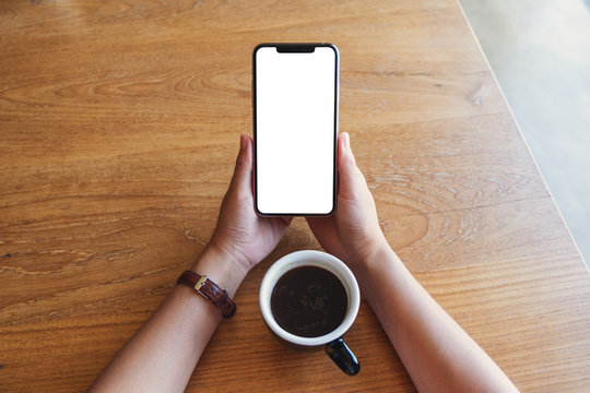 Top view mockup image of hands holding black mobile phone with blank screen with coffee cup on wooden table