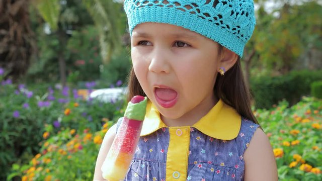 Cute little girl in a blue hat eating multicolored fruity ice cream. Trees and flowers at the background. Summer time.