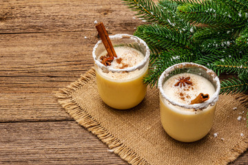 Eggnog cocktail in two glasses arranged with christmas decoration on old wooden table. Evergreen fer tree branch, artificial snow, sackcloth napkin