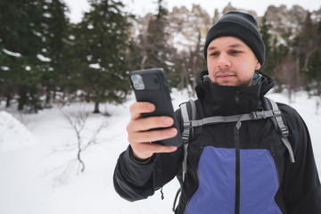 Fototapeta na wymiar Young man hiker takes photo selfie on new smartphone; high steep snowy stone slopes of mountains and pine fir forest in snowy winter day on background; tourist look at the phone; mobile navigation