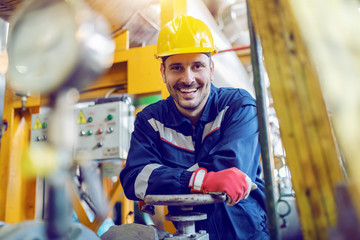 Handsome caucasian smiling unshaven worker in protective uniform leaning on valve while standing in...