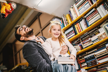 Caring father lifting his beloved only daughter wile standing in bookstore. Girl holding a book. All around are shelves with books.