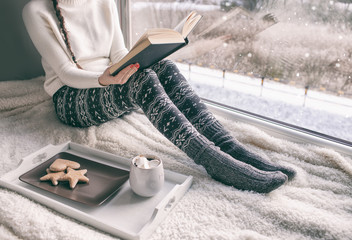 Woman sitting by the window reading book drinking coffee. Winter snowing landscape outside 