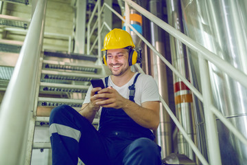 Smiling caucasian worker in overall, with helmet on head and antiphons sitting on stairs and typing...
