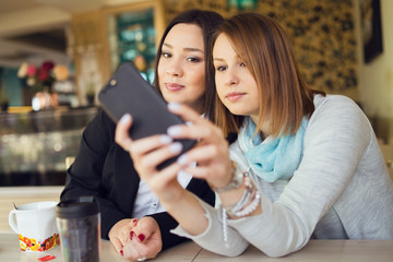 Two young women female friends using smart phone smartphone at the restaurant cafe sitting by the table video chat call or taking selfie photos photographs