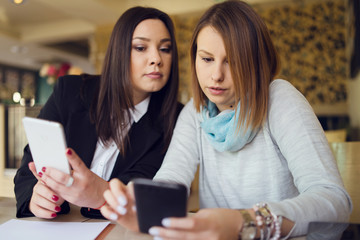 Two young women female friends using smart phone smartphone at the restaurant cafe sitting by the table reading messages or browsing internet watching video or working shopping online