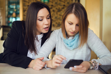 Two young women female friends using smart phone smartphone at the restaurant cafe sitting by the table reading messages or browsing internet watching video or working shopping online