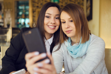 Two young women female friends using smart phone smartphone at the restaurant cafe sitting by the table video chat call or taking selfie photos photographs