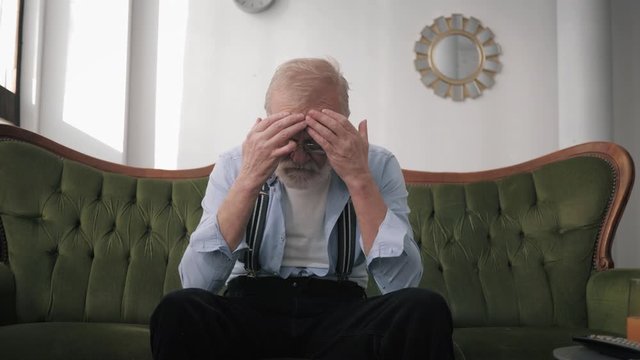 lonely elderly man with a beard and glasses for vision, suffers from depression and migraine with his hands in his head while sitting on a sofa in room