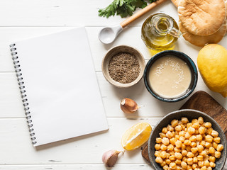 Set of ingredients for cooking homemade traditional hummus and white notebook for writing a recipe on a white wood background. Copy space.