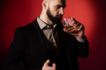 Handsome bearded man in suit with stylish beard holding a glass of whiskey and going to drink it, on red background. Close up