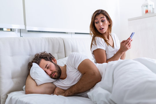 Cheating wife using mobile phone lying in bed next to his sleeping husband. Affair. Cheating Girlfriend Chatting On Phone While Boyfriend Sleeping In Bedroom At Night. Selective Focus.