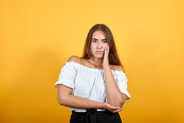 Serious young woman looking at camera, keeping hand on cheek isolated on orange background in studio in casual white shirt. People sincere emotions, lifestyle concept.