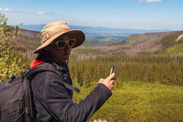 Young man tourist backpacker in travel hat standing and making photo of valley and mountains on his phone on clear sunny day. Travel landmarks, landscapes and freedom concept