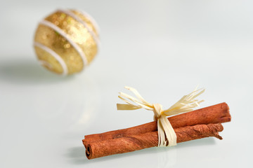 Bright still life with a close up of cinnamon sticks with a bow on a white glass plate and a golden Christmas bauble in the background