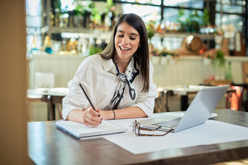 Positive attractive young businesswoman in thirties dressed in shirt sitting in cafe and writing tasks in agenda.  On table is laptop. Remote business concept.