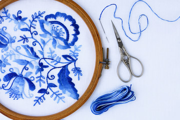 Embroidery hoop with fabric, sewing needle and thread, top view. Blue floral ornament on a white background