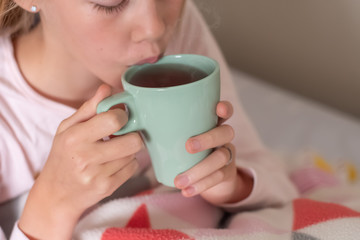 Sick teenage girl sitting on the bed covered with blanket drinking hot tea. Herbal medicine