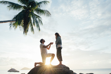 Fototapeta premium The guy makes an offer to the girl on a large stone on the seashore at sunset.
