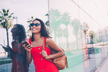Hispanic female tourist in trendy sunglasses holding modern cellular in hand and rejoicing during...