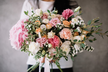 Very nice young woman holding big beautiful blossoming bouquet of fresh hydrangea, carnations, roses, eucalyptus, eustoma flowers in white and pink colors on the grey wall background