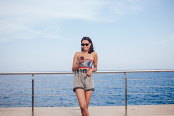 Fototapeta na wymiar Smiling millennial person in sunglasses writing content text about sea vacations during networking mobility near ocean, happy Generation Z connecting to 4g wireless during international getaway travel