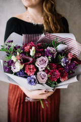 Very nice young woman holding big beautiful blossoming bouquet of fresh roses, carnations, eustoma, chrysanthemum, calla lilies flowers in pink and purple colors on the grey wall background
