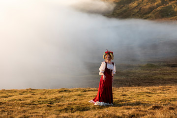 A young red-haired woman in old-fashioned clothes with a bright red skirt stays on the grass field in the highland. Faroe Islands, Denmark