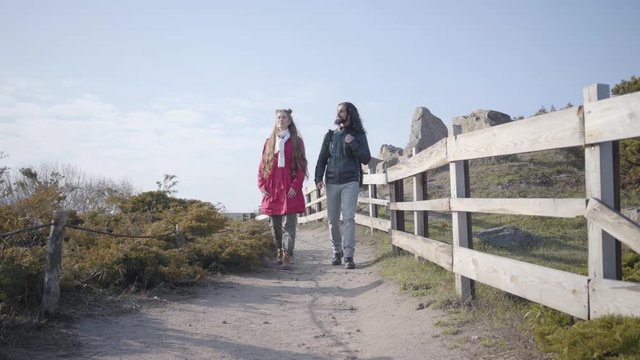 Young hippie man and woman walking along the wooden fence in autumn park. Happy biracial couple dating outdoors. Love, relationship, bonding, togetherness.