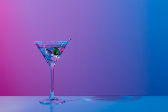 Martini cocktail with ice cubes, olive, sugar rim in neon pink and blue light on mirror bar. Night club, party, cocktail menu concept. Close-up, copy space